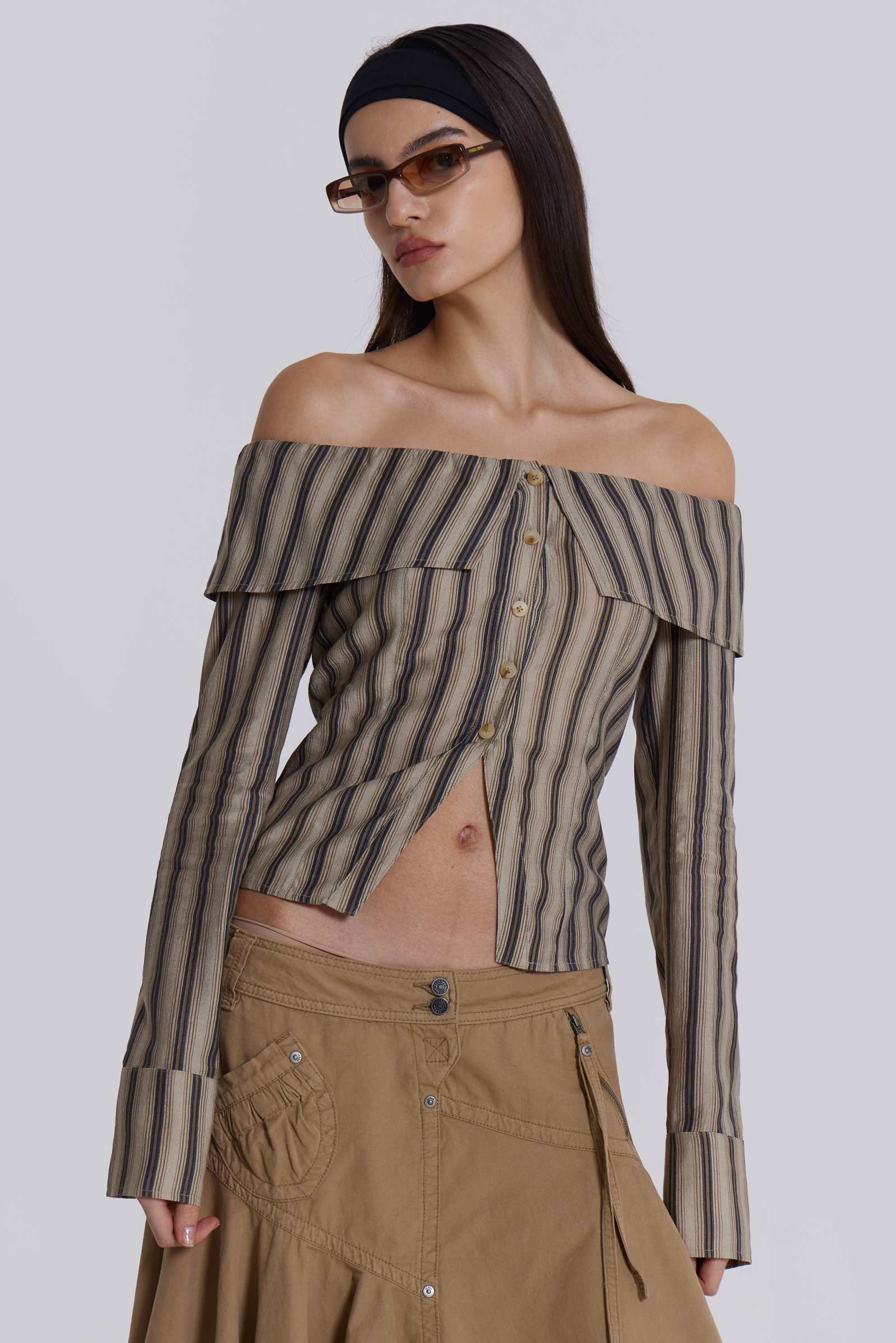  Female model wearing a khaki and brown striped off the shoulder long sleeve shirt with flared cuffs. Styled with a beige choppy cargo skirt. 