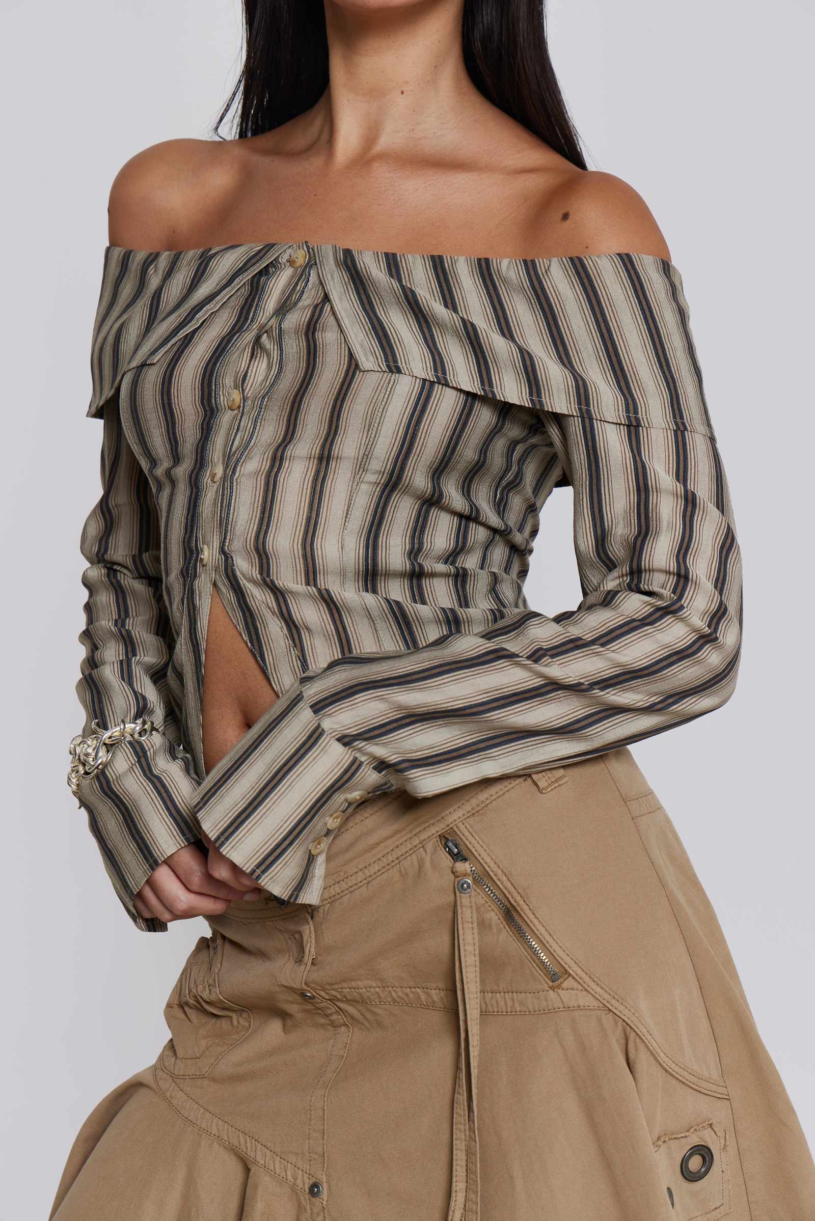  Female model wearing a khaki and brown striped off the shoulder long sleeve shirt with flared cuffs. Styled with a beige choppy cargo skirt. 