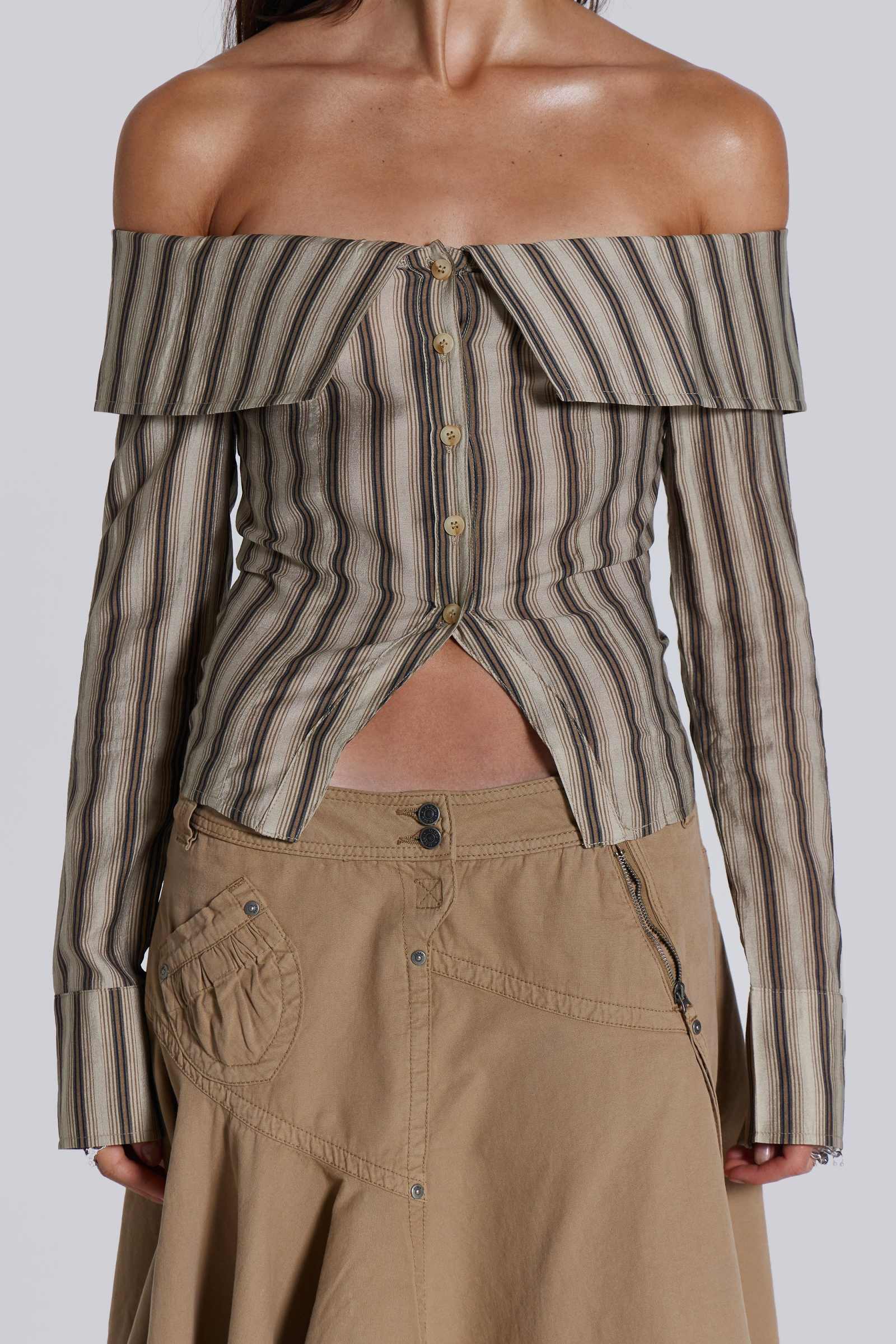  Female model wearing a khaki and brown striped off the shoulder long sleeve shirt with flared cuffs. Styled with a beige choppy cargo skirt. 