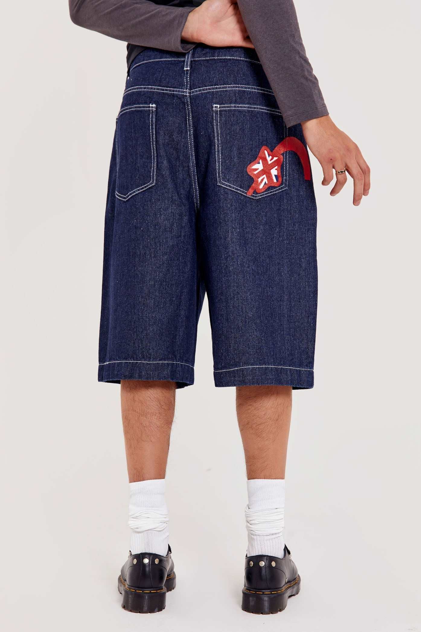 Male wearing indigo blue denim jorts in a jumbo fit with JL embroidered logo and Union Jack start branding on back pocket. 